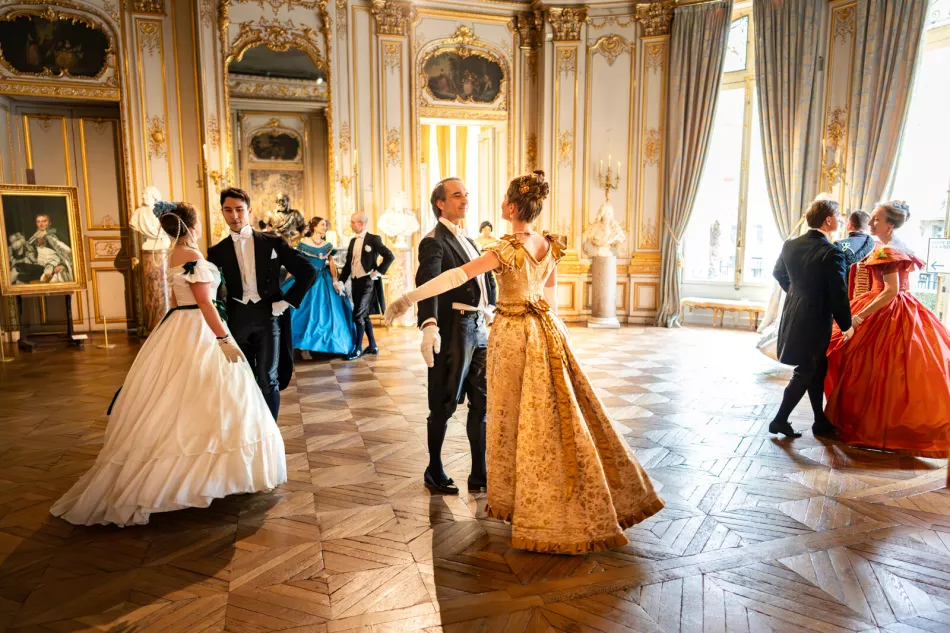 Danseurs en costumes historiques dans un salon doré, avec une danseuse en robe jaune au premier plan
